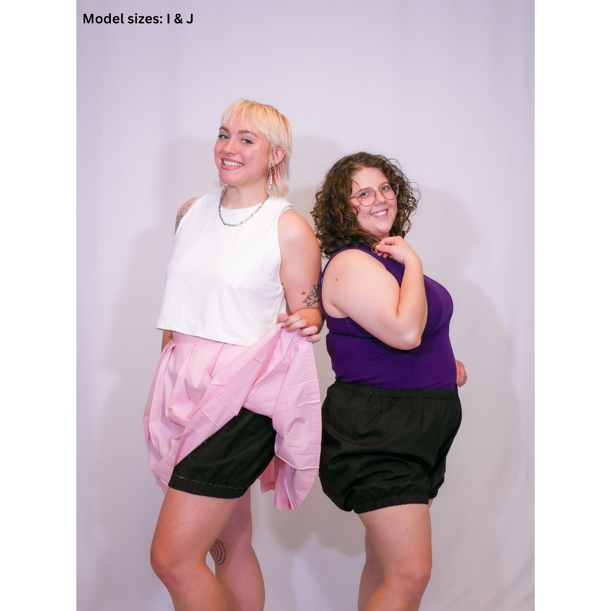 A photo of 2 women. One in a white sleeveless crop top (size H), a pink pleated skirt (size I), and short bloomers (size I). The other woman is in a purple tank top with short bloomers (Size J).
