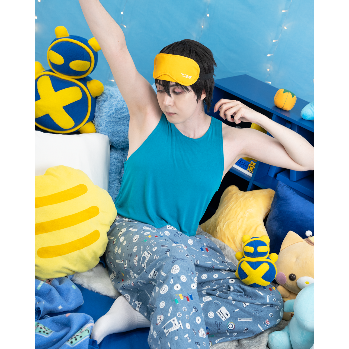 Photo of male model, matthew_aizel, wearing a sewing-themed pair of Pajama Pants and a teal crop top. He is stretching among a corner of cushions. The image elicits a feeling of sleepiness.