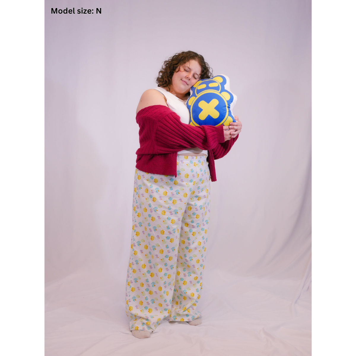 A photo of a woman wearing rubber ducky white pajama pants (size N), a white sleeveless crop top (size H), and a red jacket holding our Timmy pillow plush.