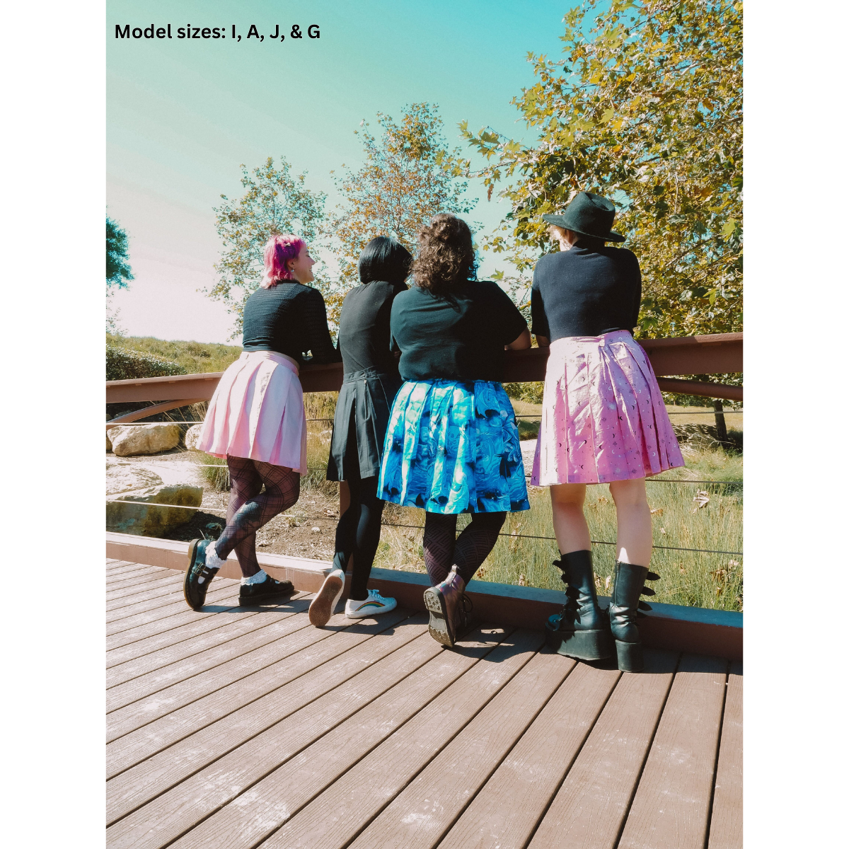 A photo of 4 people. From left to right a pink pleated skirt (size I), a black pleated skirt (size A), a blue pleated skirt with dragons (size J), and a pink starry pleated skirt (size G) all with black tops.