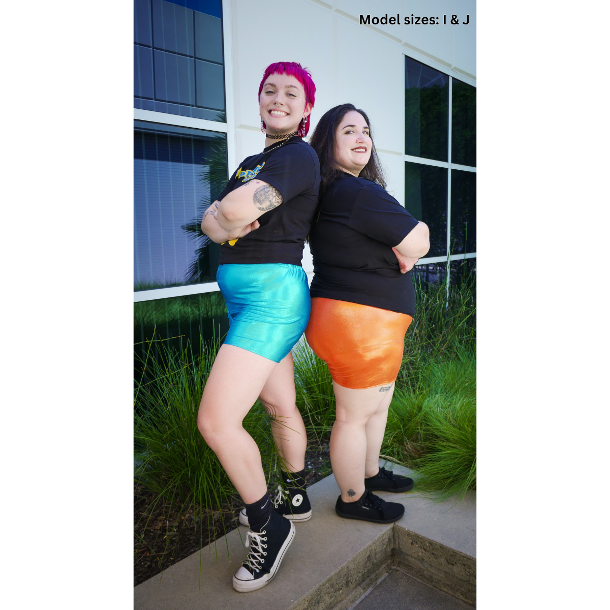 A photo of 2 people. One wearing blue Safety Shorts (size I), and the other wearing orange Safety Shorts (size J).