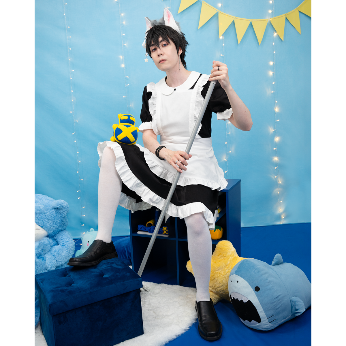 Male model, matthew_aizel, posing in the Maid Outfit (men's). He's propped on a short bookshelf and holding a broom in his hands.