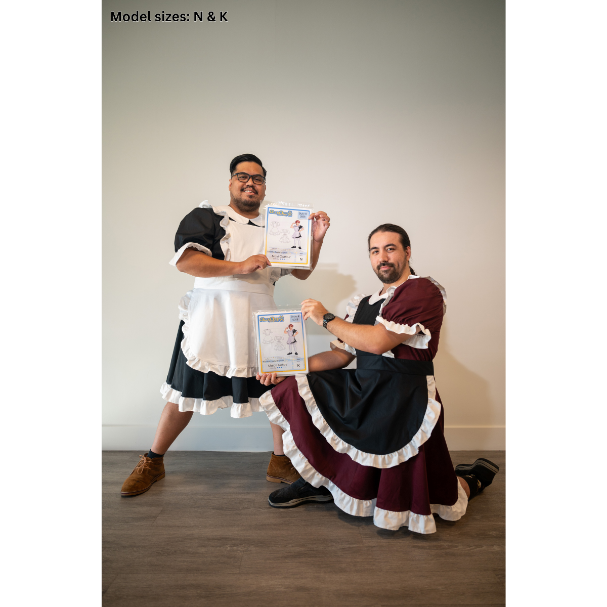 Two male models posing while wearing completed FSCO Maid Outfits (m). The model on the left is Size N, wearing a black dress with white trim and white apron. He is holding packaging for this product and smiling. The other male model is wearing a completed size K Maid Outfit. His dress is maroon with white trim, and his apron is black with white trim. He is also kneeling and holding the product's packaging.