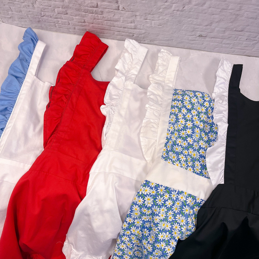 Five aprons laid out in a spread in various colors. From left to right: a white apron with blue ruffles, an apron with solid red for both the body and ruffles, a solid white apron, an apron with daisy print and white ruffles, and a black apron with white ruffles.