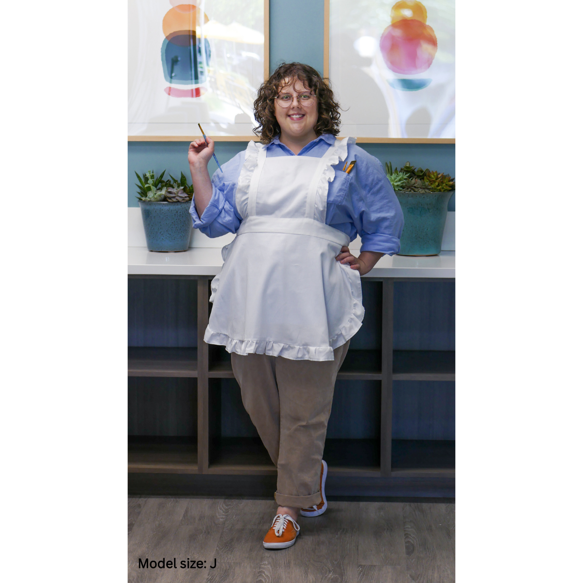 A white apron being worn by a female plus-size model. The model is facing the camera, smiling, and posing with one hand on her hip and holding a paintbrush in the other hand. The model is size J.