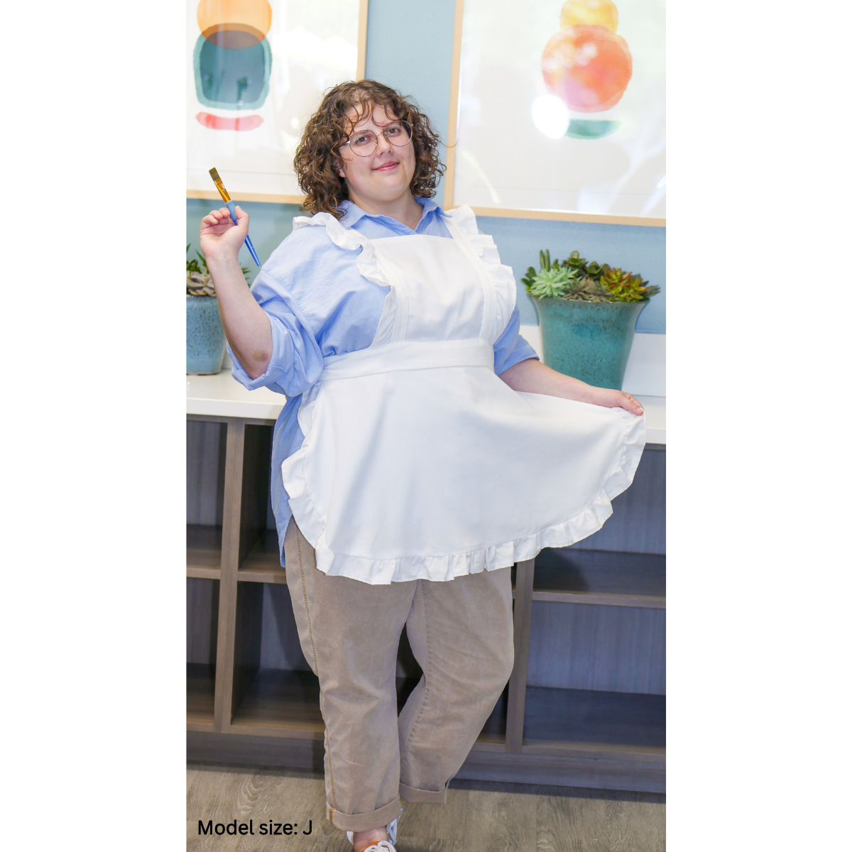 A female plus-size model posing while wearing a solid white apron. The model is holding the bottom hem of the apron with her left hand and a paintbrush in her right hand. The model is size J.