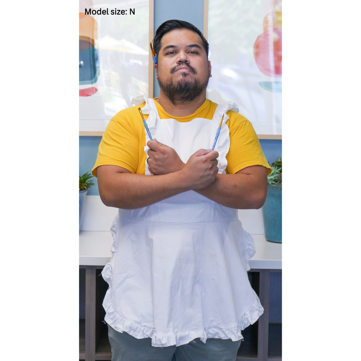 A male plus-size model posing while wearing a solid white apron. The model's arms are crossed, pointing toward his shoulders, and he is holding a paintbrush in each hand. The model is size N.