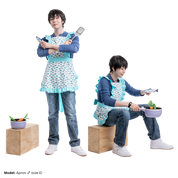 Male model, matthew_aizel, in various poses while wearing a fish-print version the Apron (men's) over a long sleeve shirt and jeans. In one variation, his arms are crossed with a fish figurine and spatula in each hand. In the other variation, he is sitting on a wooden crate and holding a pot with vegetables inside.