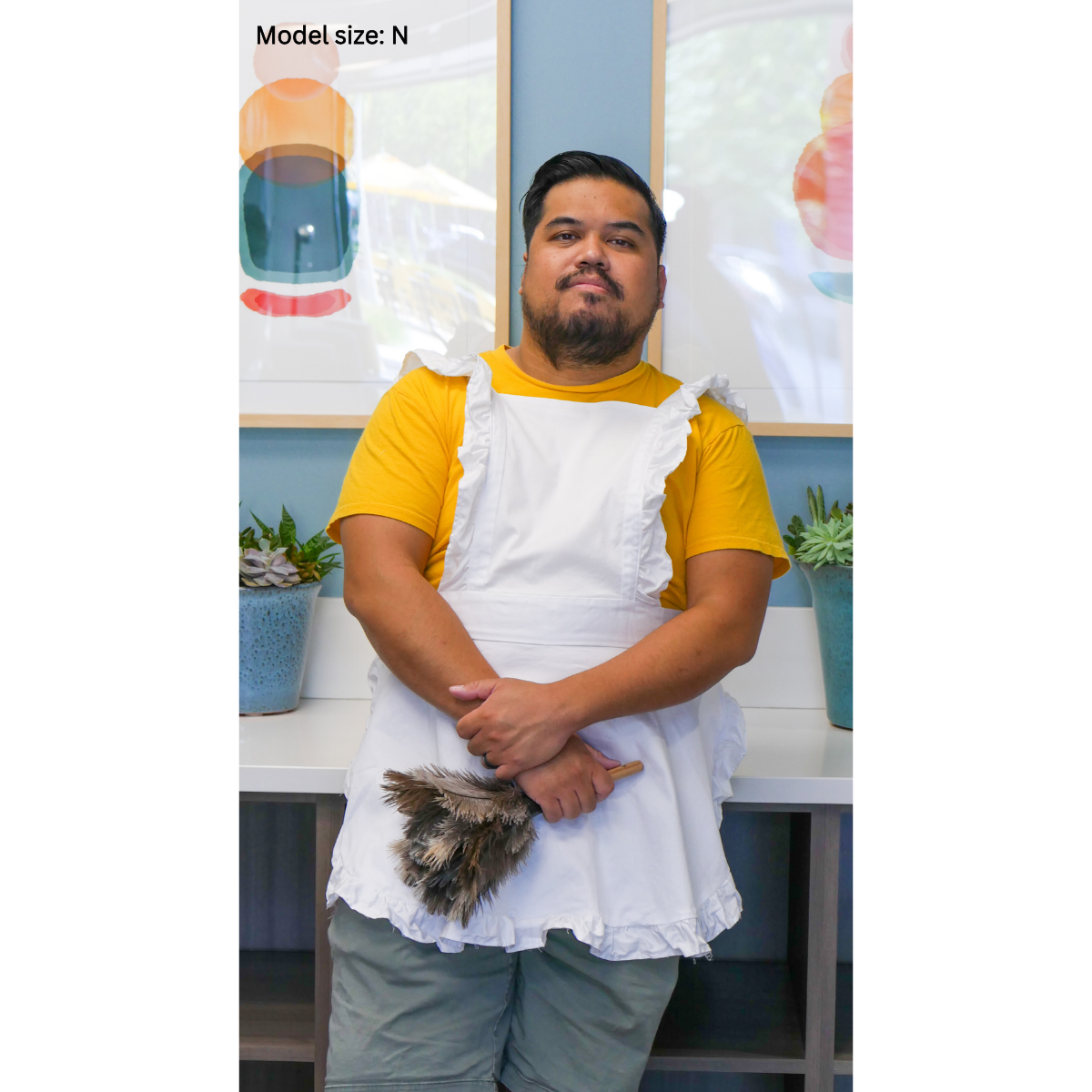 A male plus-size model modeling a white FSCO Apron (m). He is standing with a relaxed posture, and his hands are clasped in front. He's holding on to a feather duster. The model is size N.