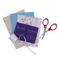 Needle Book Kit contents next to a pair of red collapsible scissors. The Needle Book kit includes fabric, glass pins in felt, a spool of thread, and ribbon.