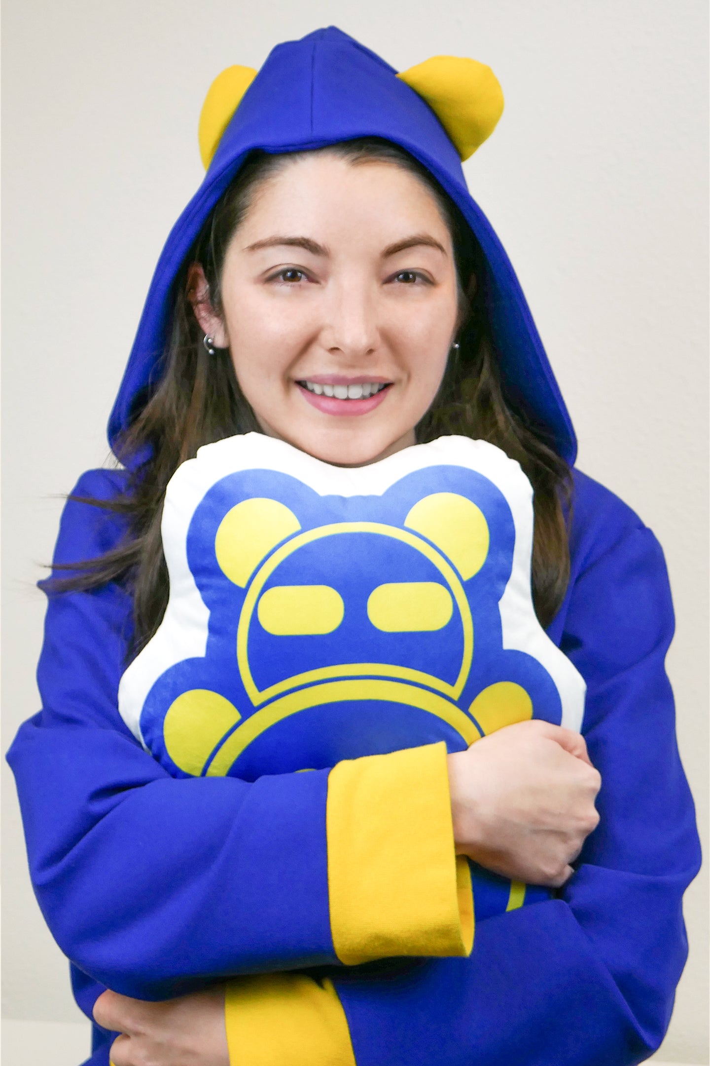 A lifestyle mage of a woman wearing a blue-and-yellow hoodie with bear ears, hugging a Timmy Pillow.