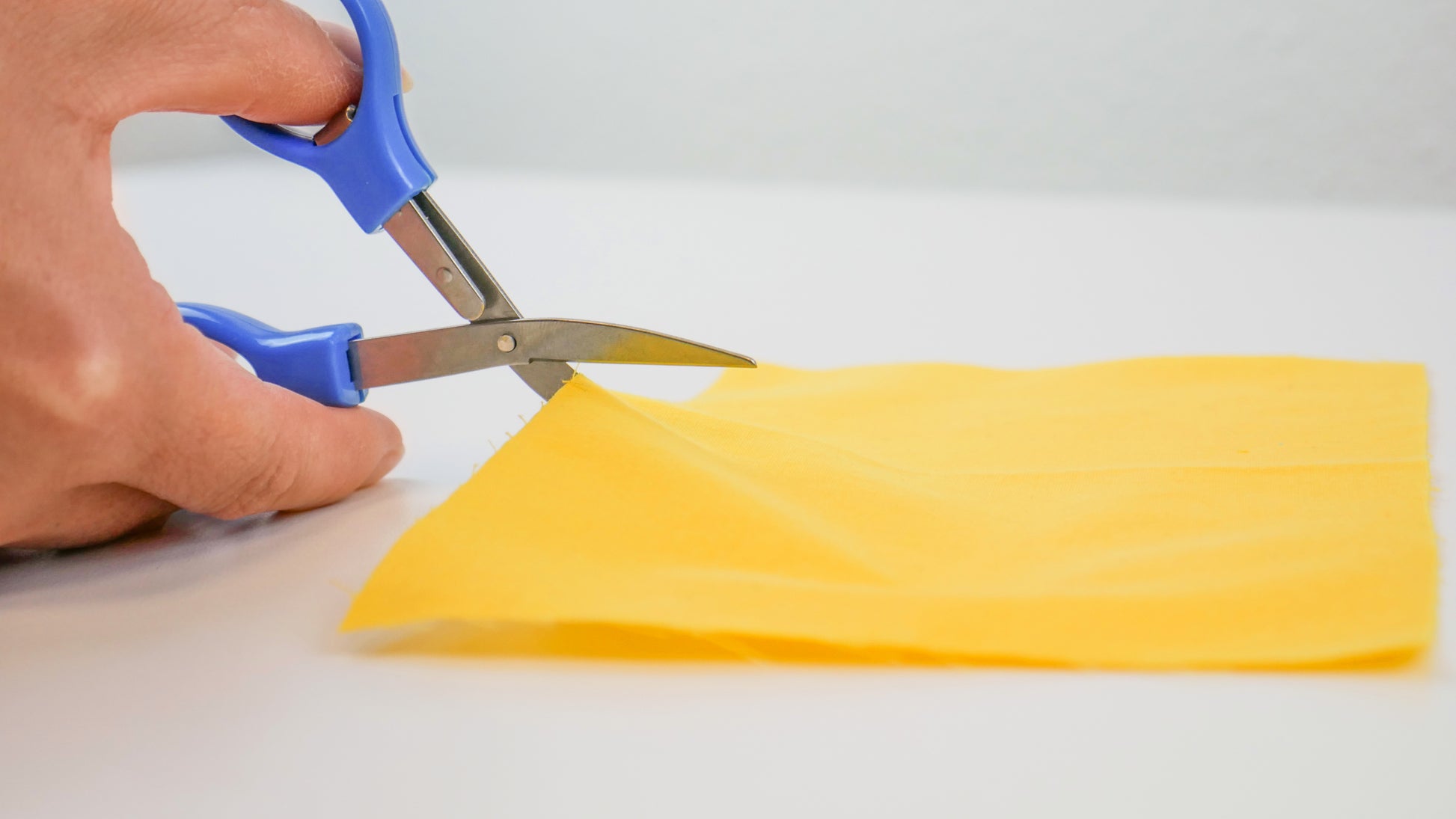 A zoomed-out shot of fingers using the blue collapsible scissors in unfolded form to cut a piece of yellow fabric.